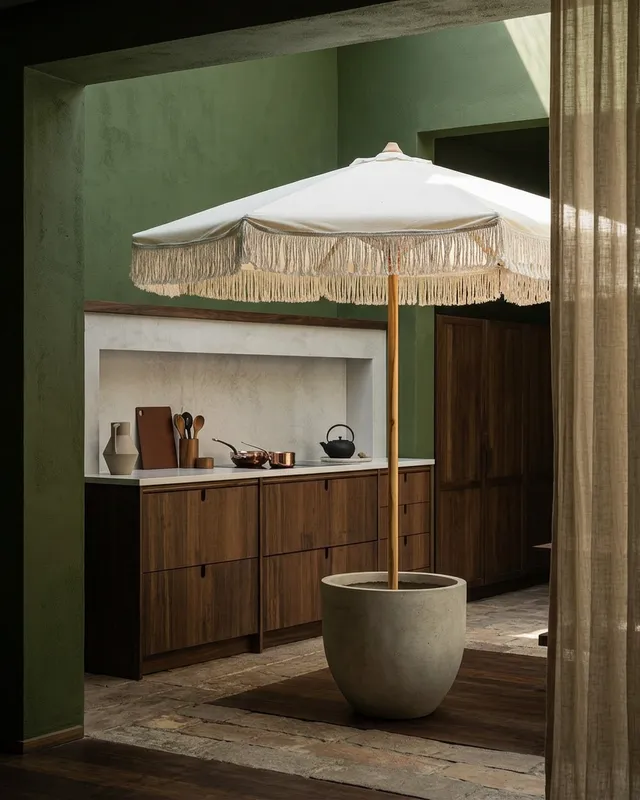 This editorial photography captures a serene indoor space featuring a large white fringed umbrella in a concrete pot, standing against a backdrop of earthy green walls and warm wooden cabinetry. The mood is tranquil and inviting, enhanced by soft, natural lighting and earthy tones.