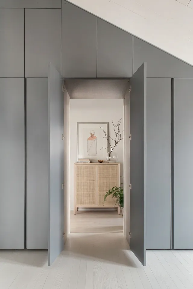 A minimalist interior scene featuring a softly lit hallway flanked by sleek gray cabinetry, leading to a light-filled room with natural wooden furniture and a framed illustration. The mood is serene and modern, dominated by neutral tones and soft textures.