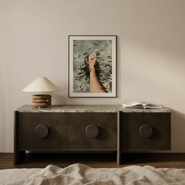 A serene interior scene featuring a wooden sideboard with a marble top, adorned with a minimal lamp and an open book, complemented by a framed photo of a foot in water. The soft, neutral tones and warm wooden hues create a calm and inviting atmosphere.
