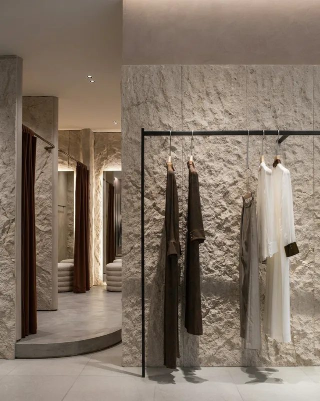 A chic retail space features earthy, textured walls with a minimalist clothing rack displaying elegant garments in muted tones. The atmosphere is upscale and serene, highlighted by soft lighting and a palette of warm browns and creamy beiges.
