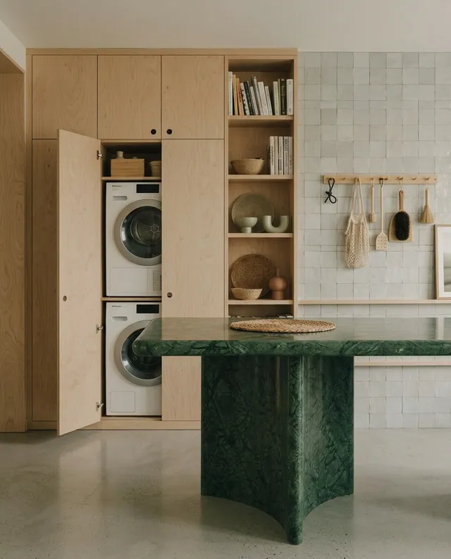 This editorial photography features a minimalist laundry room with sleek wooden cabinetry and a striking green marble table. The mood is calm and contemporary, highlighted by neutral tones and natural textures.