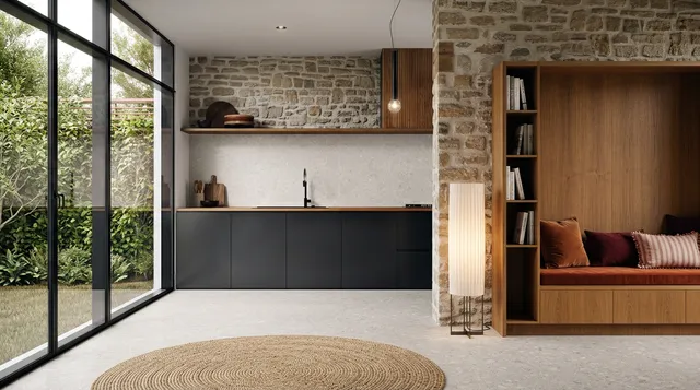 A modern interior design featuring a sleek kitchen with dark cabinetry and a cozy reading nook, all framed by natural stone walls and large glass windows. The mood is serene and inviting, with a warm color palette of browns and neutrals, complemented by greenery outside.