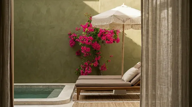 A serene editorial photograph captures a stylish lounge area beside a pool, framed by soft curtains and vibrant pink bougainvillea against a muted green wall. The warm, natural light creates a tranquil, inviting atmosphere, highlighted by earthy tones and soft textures.