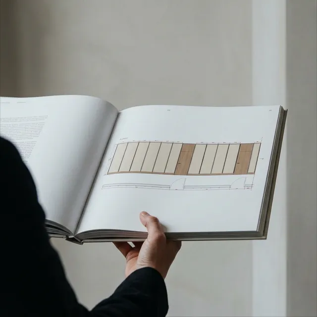 A person holds an open architectural book featuring a detailed floor plan illustration in soft earth tones. The minimalist style and muted colors evoke a calm and professional mood.