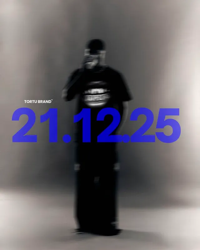 A monochromatic editorial photograph featuring a blurred figure in a black outfit, prominently displaying bold blue typography that reads "TORTU BRAND 21.12.25." The mood is modern and urban, with a focus on minimalism and striking contrast.
